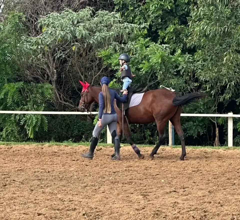 Instrutora conduz cavalo de escola com uma criança montada durante aula infantil